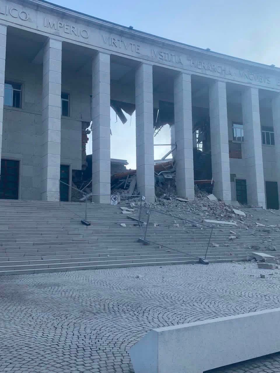 Collapsed central section of the Bolzano courthouse behind its monumental colonnade, rubble on the steps
